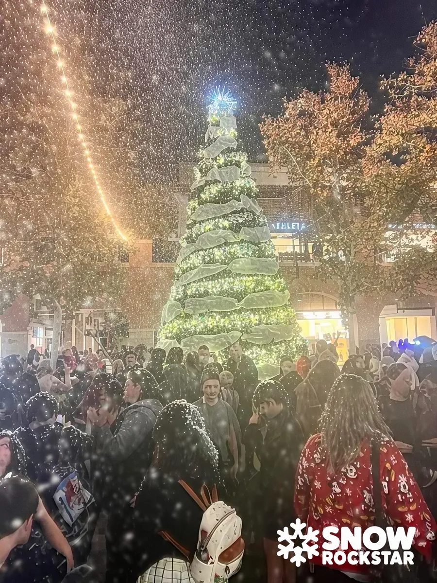 A lit holiday tree at a busy community event with heavy snow in the air and a glowing light string overhead