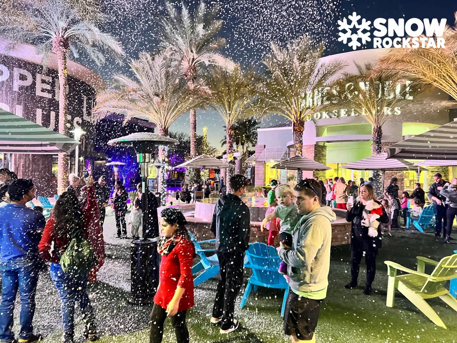 Evening crowd at a shopping plaza with palm trees, patio chairs and snow falling through the lights