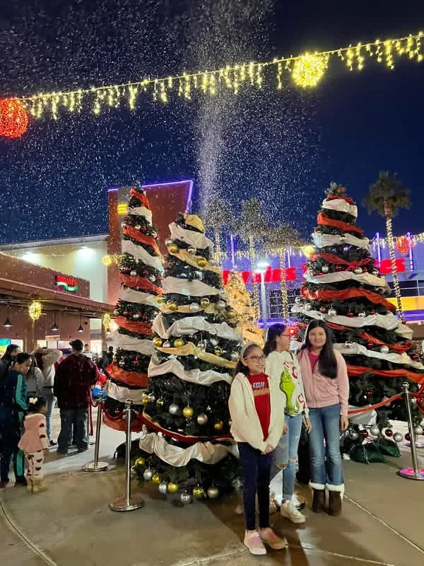 Three girls pose in front of decorated holiday trees at an evening event while snow falls behind them