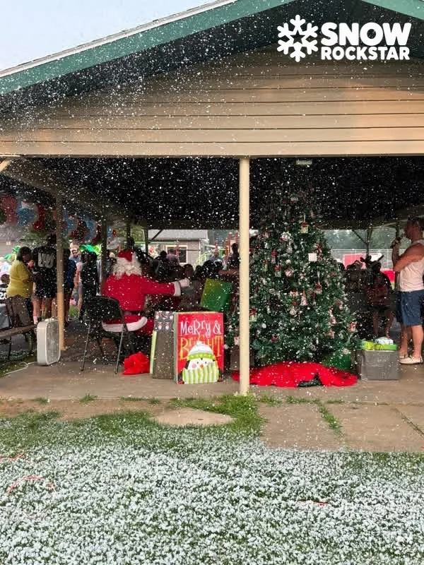 Santa seated beside a decorated Christmas tree under an open-air pavilion while snow falls over the crowd and covers the grass in front