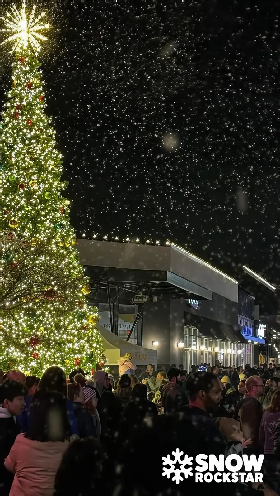 Glowing Christmas tree with a starburst topper beside a night crowd as snow falls